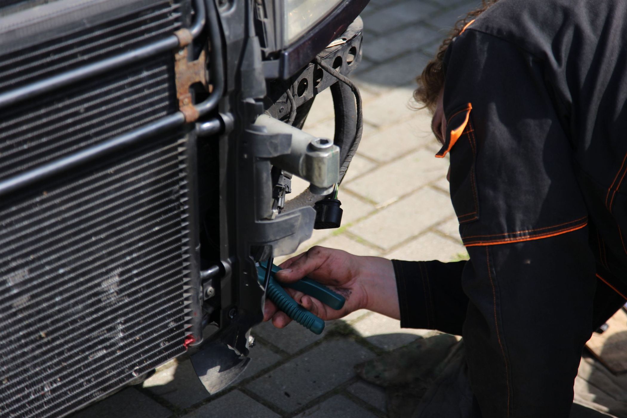 The front of a car with the bumper removed and the radiator core exposed, while someone works on the front panel.