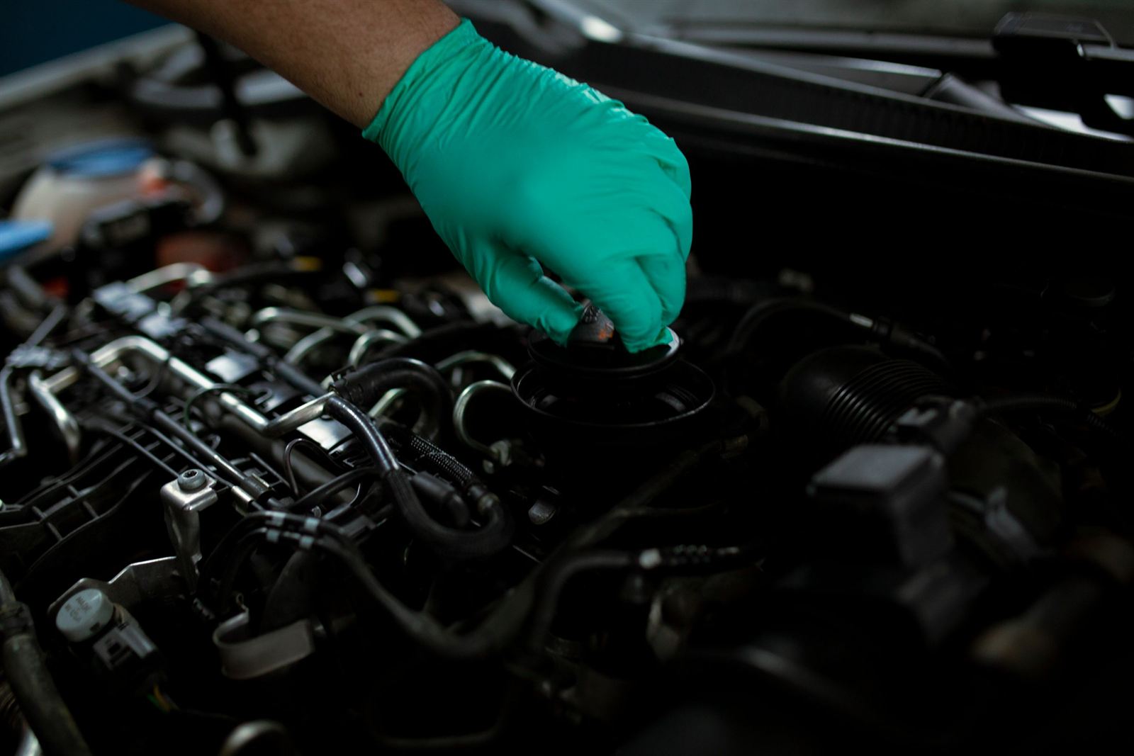 A gloved hand lifting the oil filler cap off the cam cover of a dark engine.